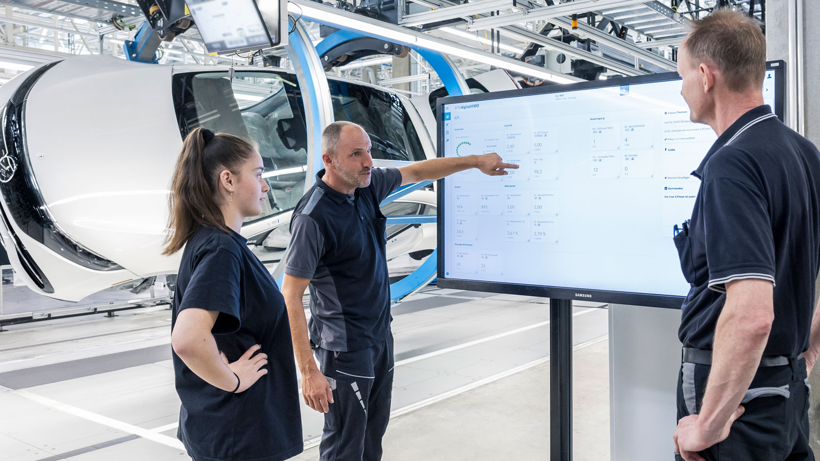 Drei Mitarbeiter der PKW-Produktion von Mercedes Benz in der Fertigungshalle vor einem Bildschirm